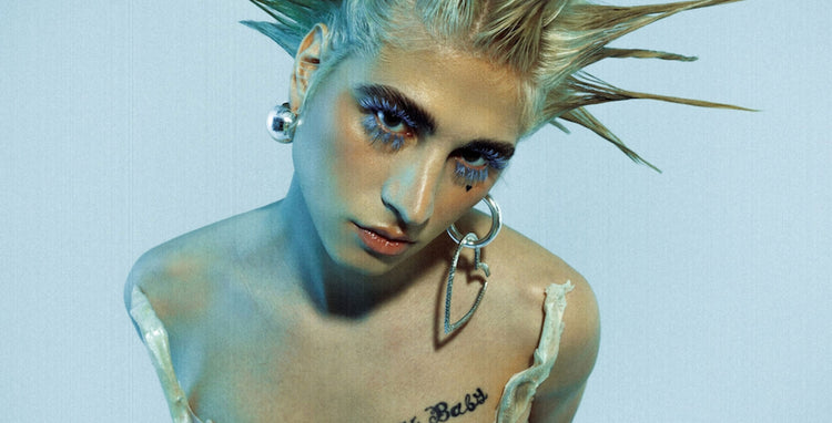 Edgy model with spiked blonde hair, bold blue lashes, and chest tattoo - a fearless look that reflects tattoo culture and the empowered, pain-free experience championed by Tattoo Numbing Cream Co.